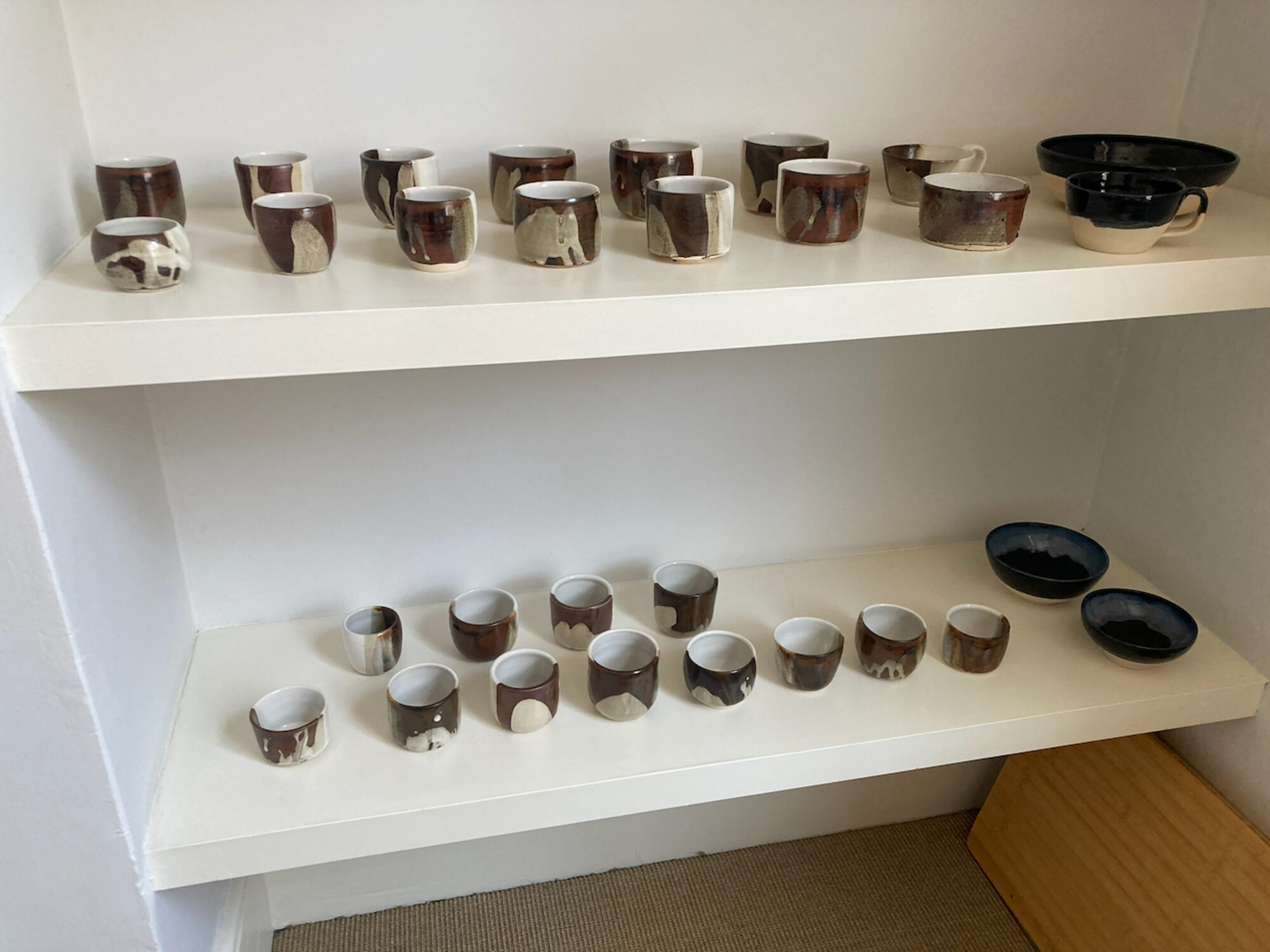 Rows of handmade cups, tumblers and bowls in brown and cream glazes, arranged on white shelves