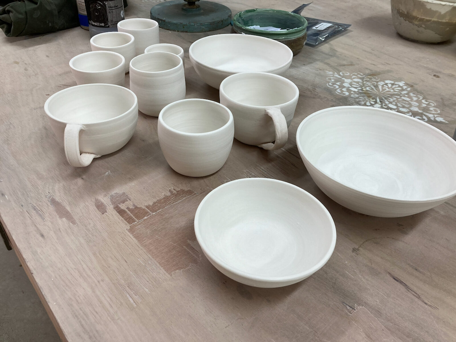 Unglazed bisque-fired cups and bowls lined up on a workshop table, ready for glazing